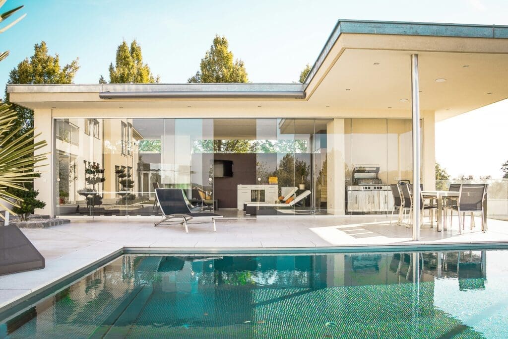Modern house with large glass walls, outdoor lounge chairs, a dining area, and a pool in the foreground. The setting is bright and sunny, surrounded by trees.