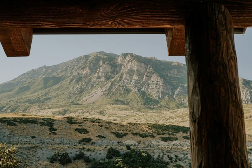 View of a rugged mountain with green and brown slopes, seen from under a wooden structure with beams and a post in the foreground. The sky is clear and blue.