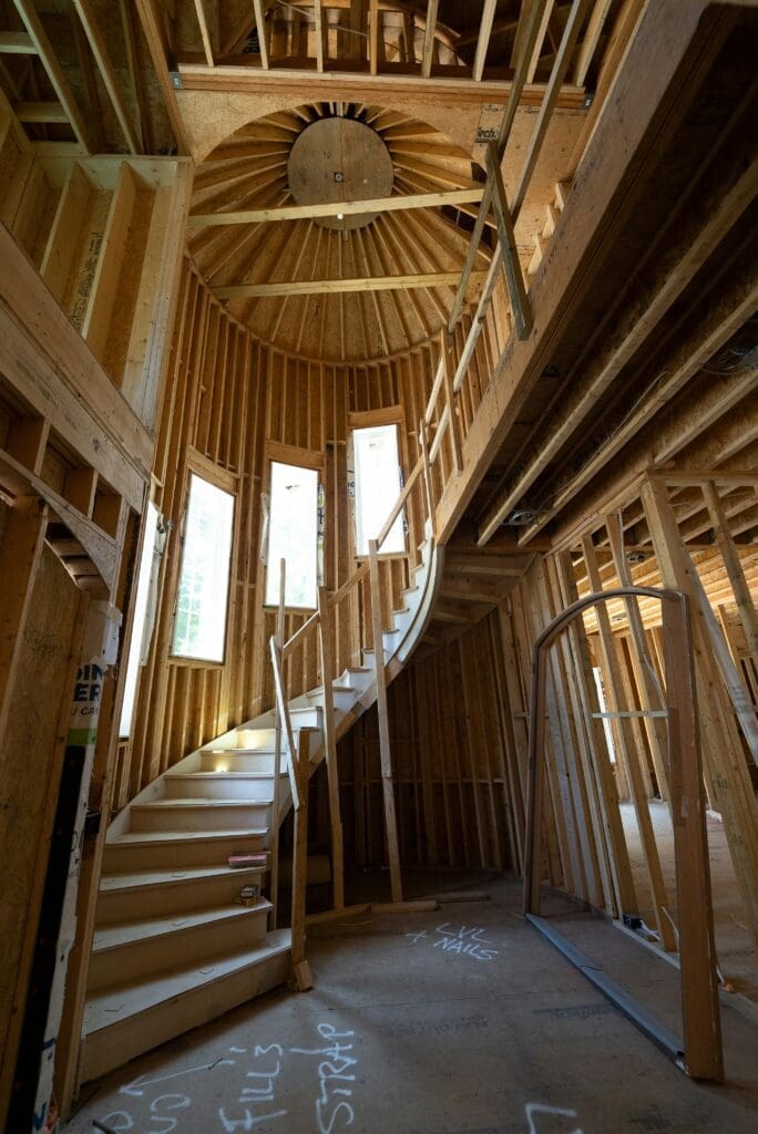 A curved staircase in a house under construction, surrounded by exposed wooden framing. Three tall windows let in natural light, and a circular ceiling structure is visible above the stairs.